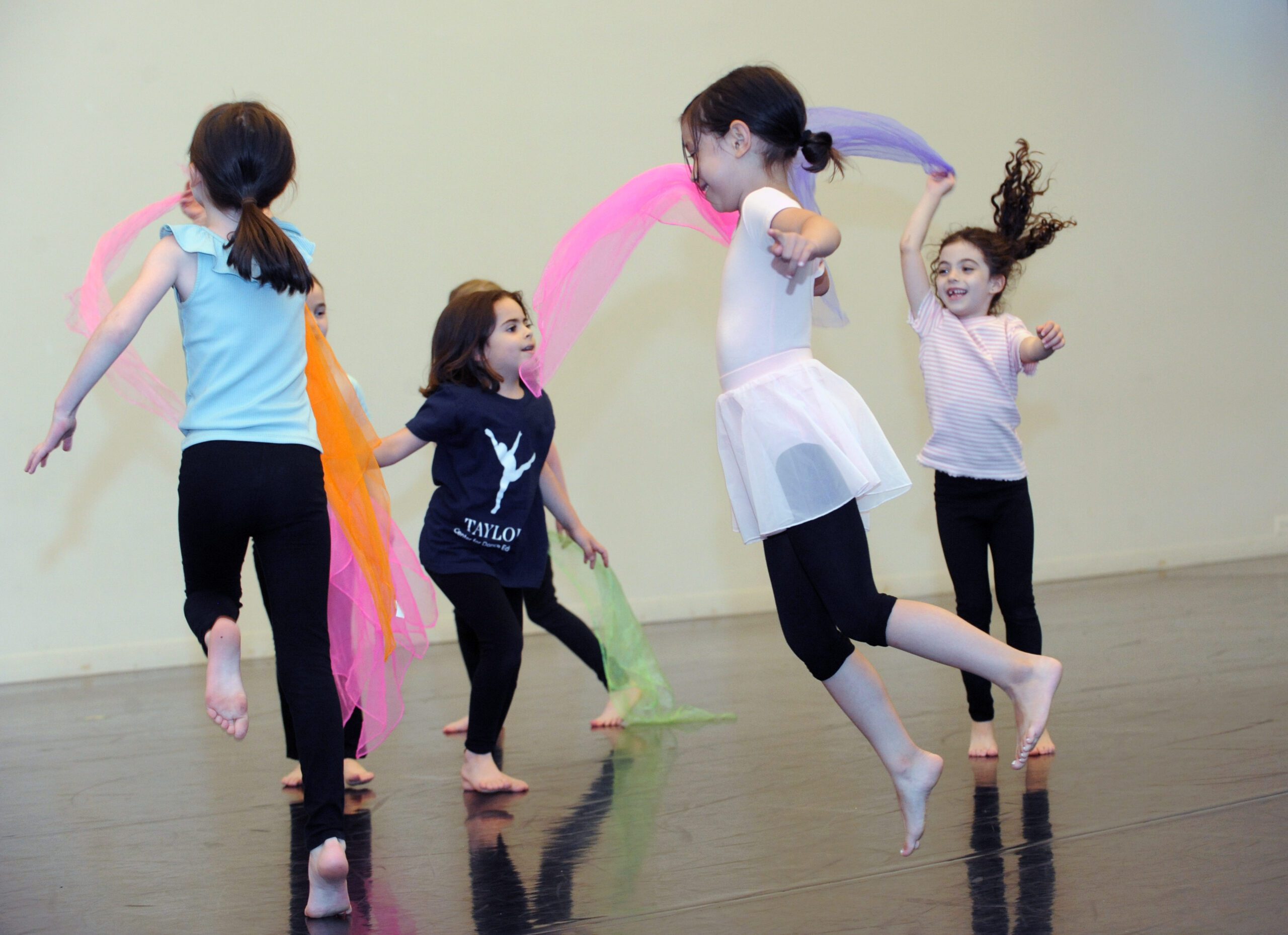 Four young dancers jumping and moving through a Taylor School dance class with colorful scarves